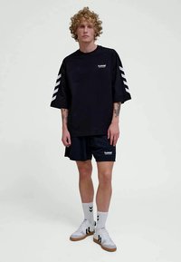 Ungdom med krøllete hår iført svart, oversized sportstøy, hvite sokker og hvite joggesko foran en ensfarget lys bakgrunn.