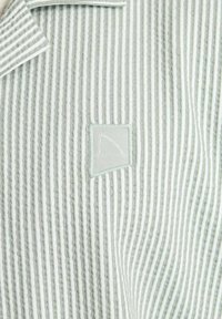 Chemise collerette à rayures verticales vertes et blanches. Dotée d'un petit patch carré avec un design brodé. Fabriquée en tissu texturé.