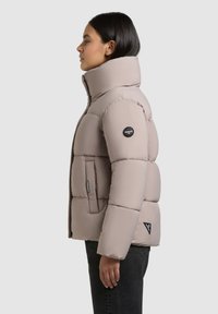 Puffer jakke i blød beige, med høj krave, store kviltede sektioner, sidelommer og et logo-patch på ærmet.