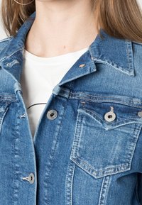 Vaaleansininen farkkutakki, jossa on terävä kaulus, kaksi rintataskua, hopeanväriset napit ja näkyvät ompelutiedot. Sileä rakenne.