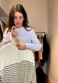 Kvinde holder en hvid sweater med sorte vandrette striber på en bøjle inde i et rum med tøj, der hænger på et stativ bag hende.