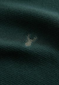 Una tela de punto en verde oscuro presenta un sutil logo de ciervo bordado en gris en la superficie, con una apariencia fina y texturizada.