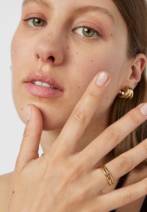 Unas pendientes de aro dorados y un anillo dorado son llevados en la mano de una persona de piel clara, con una superficie lisa y pulida y diseños curvados.