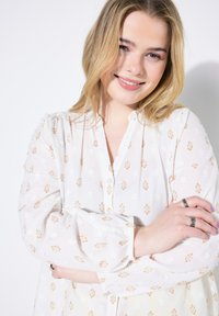 Blusa bianca con motivo floreale in tonalità arancioni e gialle, tessuto con texture, chiusura frontale con bottoni e maniche lunghe con polsi arricciati.