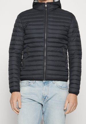 Homme portant une veste matelassée noire à capuche avec des coutures horizontales et un jean bleu clair, debout devant un fond uni.