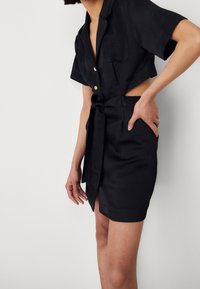 Robe courte noire en tissu lisse, avec un design court, des manches courtes, une poche poitrine et un détail de taille nouée. Boutons dorés présents.