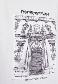 Bílý tričko s černým náčrtem architektonické fasády s výraznými dveřmi, nahoře s textem "EMPORIO ARMANI".