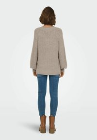 Pull tricot beige clair avec un motif texturé, manches longues, et une longueur d'ourlet arrière légèrement plus longue, associé à un jean skinny bleu et des bottines marron.