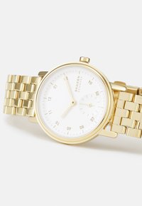 Zlaté kovové náramkové hodinky s kulatým bílým ciferníkem, hodinovými značkami, dvěma ručičkami a designem náramku z pletiva. Minimalistický styl s čistými liniemi.