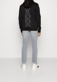 Sudadera negra con un gran logo blanco en la parte posterior, combinada con jeans de un azul claro y zapatillas blancas con rayas rojas.