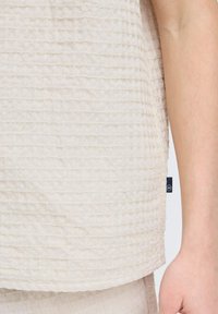 Top texturizado blanco roto con un patrón arrugado, con un cuello redondo y una pequeña etiqueta de la marca en el costado, combinado con una parte inferior de color similar.