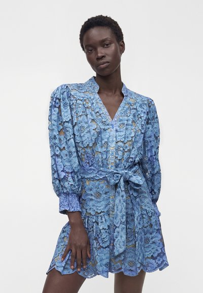 Femme portant une robe courte en dentelle bleue à manches longues avec un motif floral et une ceinture nouée à la taille, debout devant un fond blanc uni.