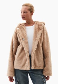 Beige umělá kožešinová bunda s kapucí, velkými klopami, knoflíkovými zapínáními a dlouhými rukávy, nošená přes bílou košili. Měkká textura a neformální design.