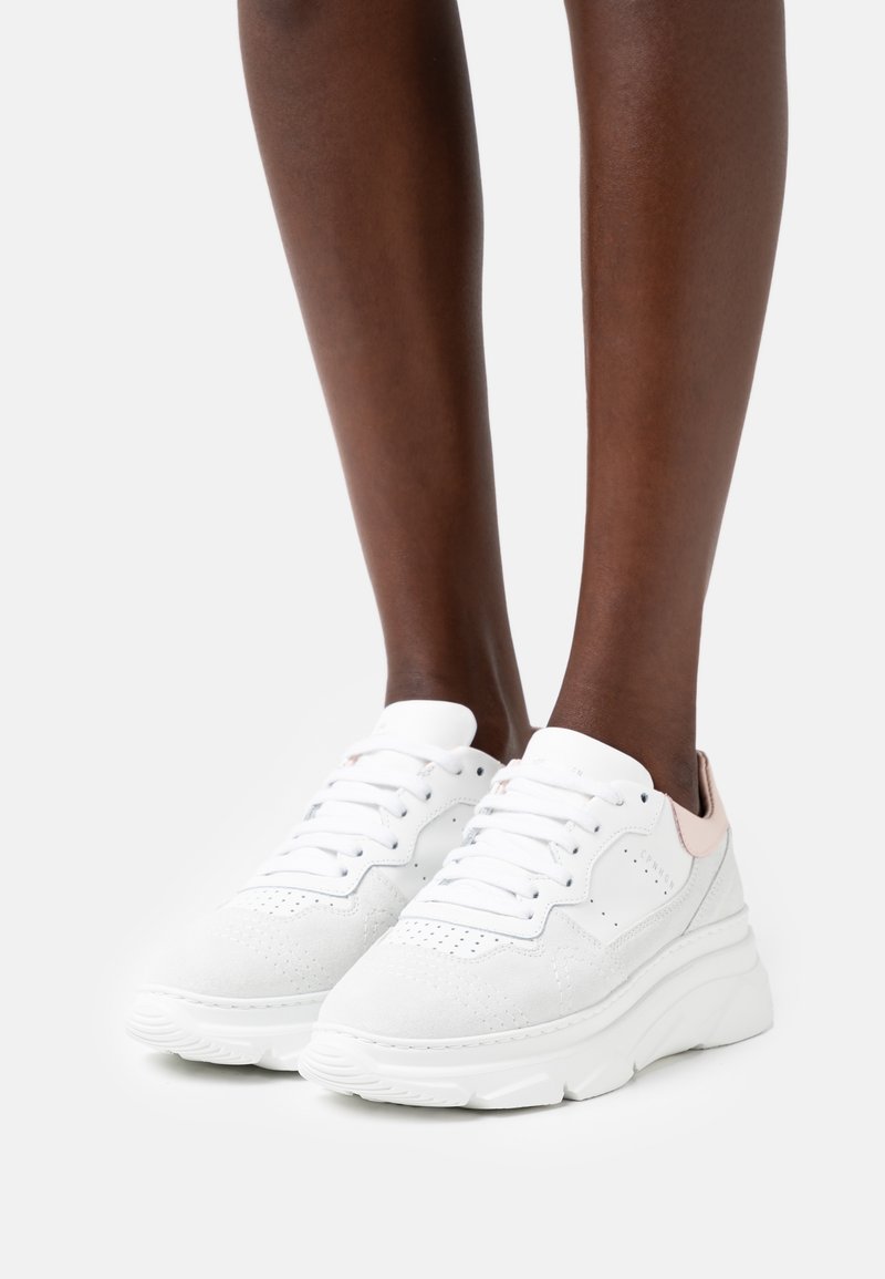 Baskets à plateforme blanches avec un design à lacets portées sur des jambes à peau foncée, présentant des détails perforés et un accent talon rose clair.