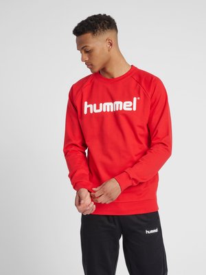 Man die zijn mouw rechtzet, draagt een rode Hummel trui en zwarte Hummel broek tegen een effen achtergrond.