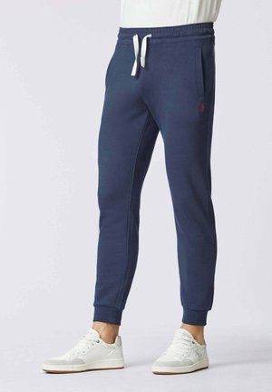 Uomo che indossa pantaloni da jogging blu navy con coulisse bianca e sneakers bianche, in piedi contro uno sfondo chiaro uniforme.