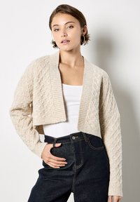 Cárdigan de punto en beige con un patrón texturizado, longitud cropped y abertura frontal. Combinado con un top blanco y jeans de talle alto en azul oscuro.