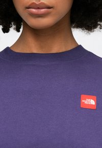 Felpa viola con collo a costine. Presenta un piccolo logo quadrato rosso di "The North Face" sulla parte anteriore. Texture in cotone morbido.