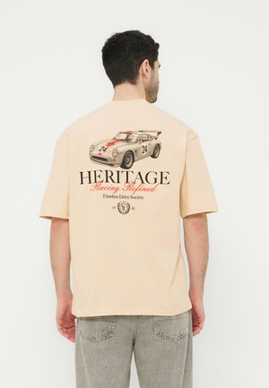 Férfi bézs pólóban, melyen a hátoldalon vintage versenyautó grafika, 24-es szám és a "HERITAGE Racing Refined Timeless Drive Society" felirat látható.