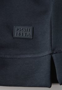 Tejido de punto azul marino con un parche rectangular de logotipo texturizado. Presenta una abertura lateral y costuras prolijas. Material suave y elástico.