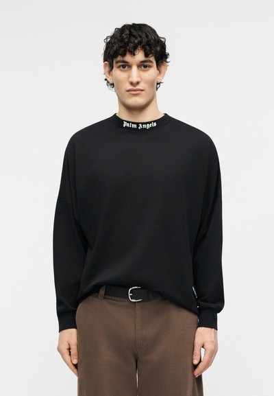 Giovane uomo con capelli ricci indossa una maglia nera a maniche lunghe con la scritta "Palm Angels" sul colletto e pantaloni marroni su sfondo semplice.
