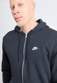Sötétszürke, cipzáras kapucnis pulóver, pamut keverékből készült; fehér Nike logó található a mellrészen, húzózsinórok és elülső zseb jellemzi.