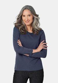 Langarm-Navy-Hemd aus weichem Material mit Rundhalsausschnitt, kombiniert mit dunklen Jeans; getragen von einer Frau mit langen, welligen grauen Haaren.