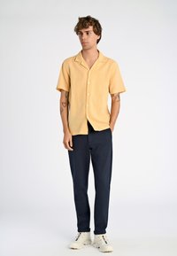 Giovane con capelli ricci che indossa una camicia gialla a maniche corte, pantaloni blu navy e sneakers bianche, in piedi con le mani parzialmente nelle tasche.