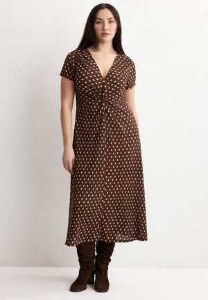 Žena v hnedom krátkom šatom s výstrihom do V a svetloružovými bodkami, obutá v hnedej semišovej obuvi až po kolená, stojaca na jednoduchom pozadí.