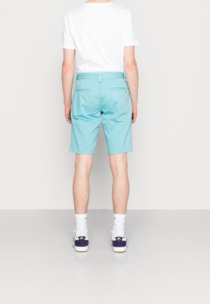 Persona vistiendo pantalones cortos color teal hasta la rodilla, camiseta blanca de manga corta, calcetines blancos y zapatillas Vans, de pie frente a un fondo liso.