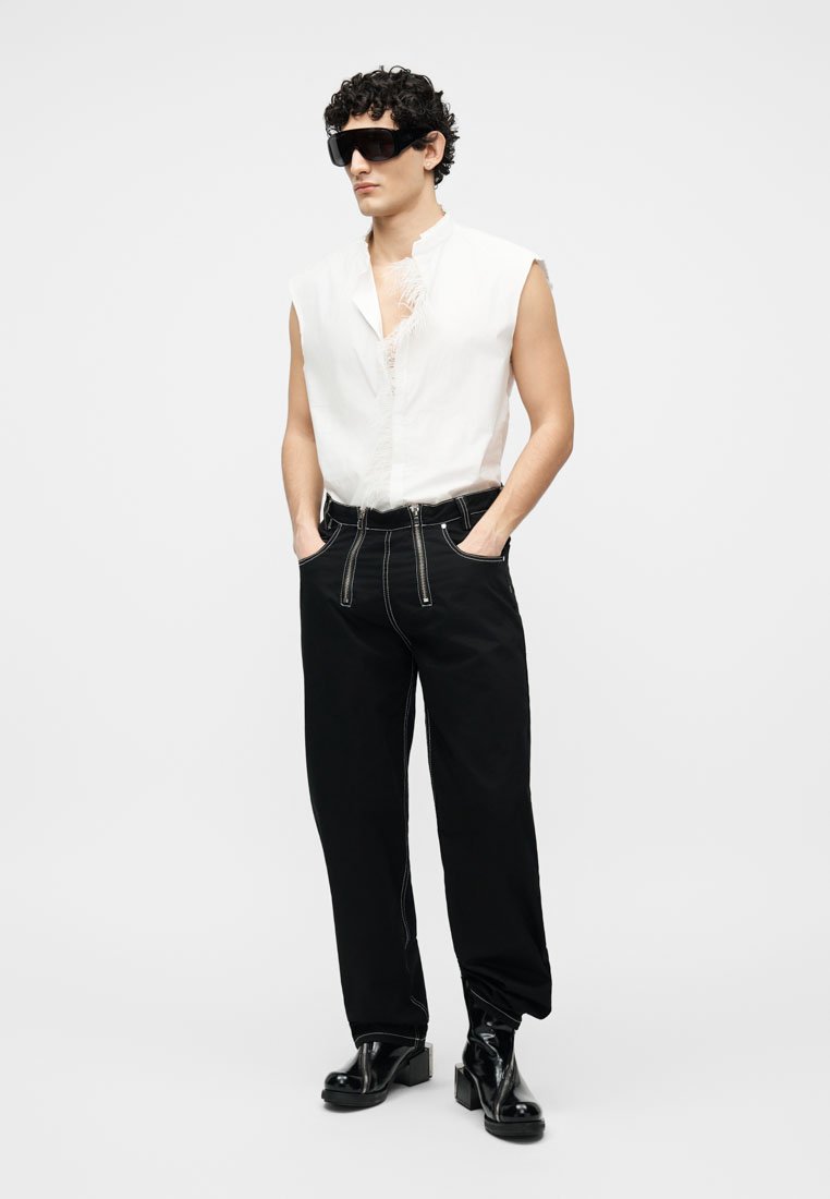 Homme portant un débardeur blanc, un pantalon noir avec des coutures contrastantes, des bottes plateformes noires, et de grandes lunettes de soleil noires, debout les mains dans les poches.