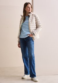 Doudoune beige clair avec zip, accents verts ; pull rayé bleu et blanc ; jeans bleus ; baskets blanches ; debout contre un fond neutre.