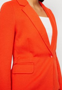 Blazer arancione brillante con un tessuto testurizzato, rever a punta, chiusura con un solo bottone e dettagli con tasche laterali.