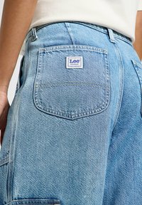 I jeans in denim blu chiaro presentano una tasca posteriore con un'etichetta del logo Lee Workwear bianca e blu. La trama mostra un classico motivo a tessitura denim.