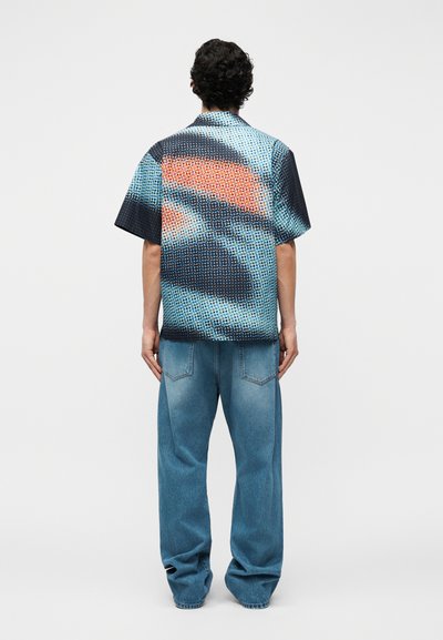 Homme portant une chemise à manches courtes à boutons avec un motif abstrait bleu, noir et orange, associée à un jean bleu ample, debout de dos.