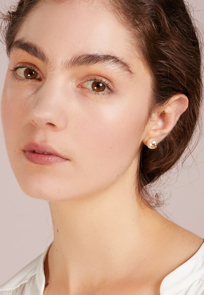 Orecchini a bottone in oro con perla al centro, forma rotonda, superficie liscia e design minimalista, indossati da una persona con occhi castani.