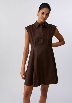 Donna che indossa un vestito senza maniche di colore marrone scuro con colletto e gonna svasata, in piedi con un braccio piegato dietro la schiena.