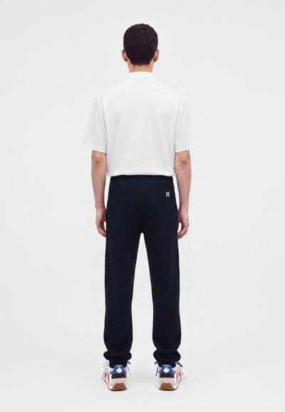 Homme debout de dos, portant une chemise blanche à manches courtes, un pantalon noir avec un petit logo blanc et des baskets blanches avec des détails bleus.