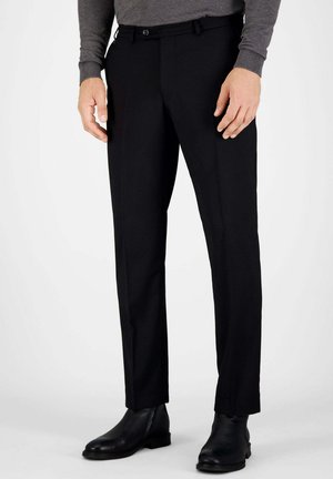 Mann trägt schwarze Anzughose, schwarze Lederknöchelstiefel und graues langärmeliges Oberteil, steht vor einfarbigem Hintergrund.