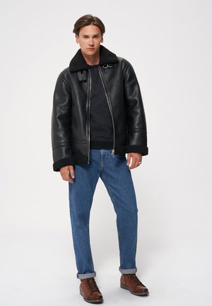 Czarna skórzana kurtka z kołnierzem ze sztucznego futra, srebrnymi zamkami i brązowymi akcentami, zestawiona z teksturowanym granatowym swetrem i niebieskimi dżinsami.