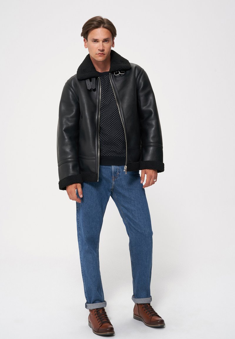 Czarna skórzana kurtka z kołnierzem ze sztucznego futra, srebrnymi zamkami i brązowymi akcentami, zestawiona z teksturowanym granatowym swetrem i niebieskimi dżinsami.