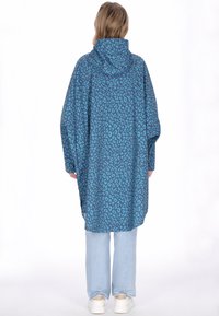 Túlméretezett kapucnis esőkabát tengerzöld színben fekete leopárd mintákkal, laza ujjak és egyenes szabás jellemzi, világoskék farmer és fehér cipő fölött viselve.