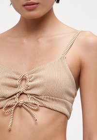 Stripet bikini topp i beige og hvit, med en samlet frontdesign med en nøkkelhullsdetalj og justerbare knytinger i bunnen.
