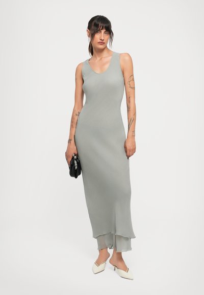 Woman with tattoos wearing a sleeveless light gray maxi dress, holding a black clutch, and white pointed mules against a plain background.