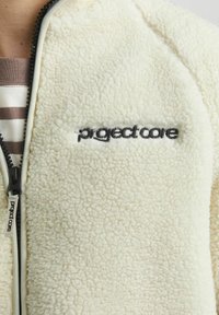 Beežikas fliisk jacket tekstuuri pinnaga, musta tõmbluku, kontrastse sisemise voodriga ning esiküljel tikitud logoga.
