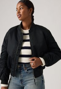 Donna che indossa un bomber nero sopra un maglione a righe bianche e blu navy e jeans blu, guarda di lato con i capelli intrecciati.