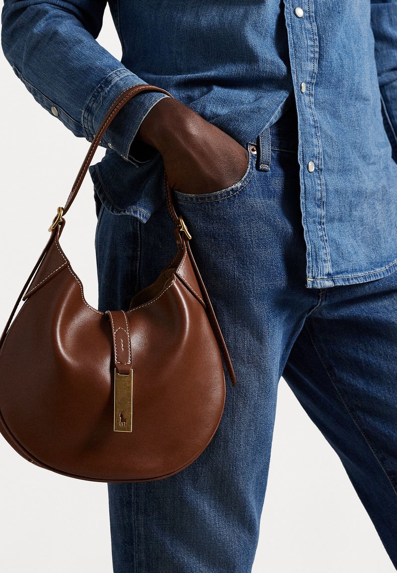 Rjava usnjena hobo torba zaobljene oblike, ki vsebuje zlatokovinske dodatke in kontrastno šivanje. V kombinaciji z denim srajco in kavbojkami.