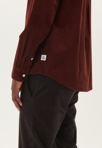 Bordeaux corduroy overhemd met een puntkraag, knopenmanchetten en een genaaid label met maat. Gecombineerd met donkere broek, zijaanzicht zichtbaar.
