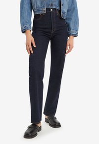 Personne portant un jean bleu foncé taille haute à jambes droites, des chaussures noires sans lacets et une veste courte en jean bleu sur fond blanc.