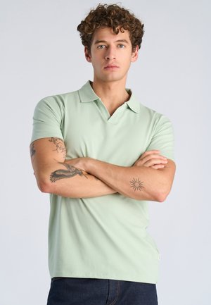 Giovane uomo con capelli ricci e occhi azzurri, indossa una polo verde chiaro, braccia incrociate che mostrano tatuaggi su entrambi gli avambracci.